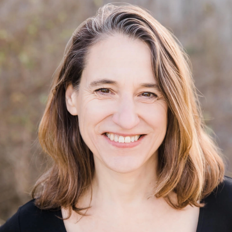 Amanda Quaid, a woman with light skin and straight, shoulder-length brown hair, smiles warmly at the camera. She wears a black top and stands outdoors with a blurred background of greenery and light brown tones.