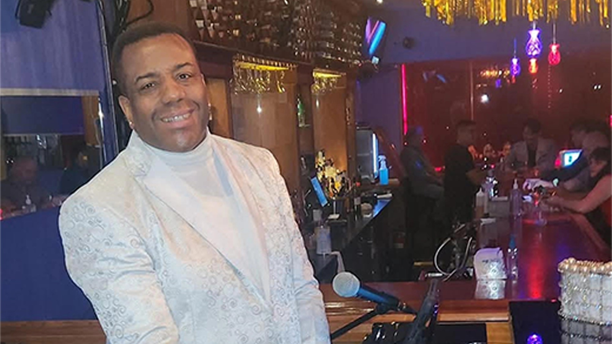 Kenny Allan Smith site image 26 A man wearing a white patterned blazer and turtleneck smiles while standing beside a piano in a lively bar with colorful lights, a drinks counter, and patrons—setting the scene for Kenny Allan Smith at Kenny’s Turn.