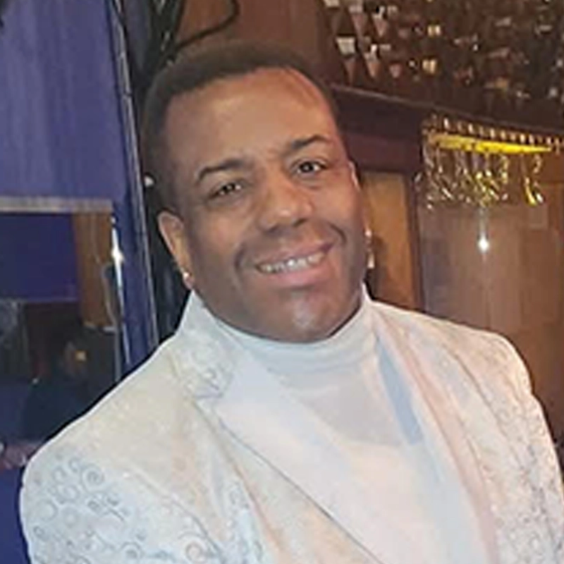 A man with short hair smiles at the camera. He is wearing a light-colored, patterned suit jacket over a white turtleneck. The background features reflective surfaces and blue and gold tones, suggesting an indoor, possibly formal, setting.