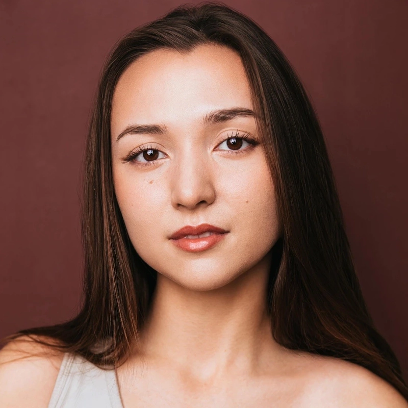 A young woman with straight, long brown hair poses against a maroon background. She has light skin, full lips with natural lipstick, and minimal eye makeup. Jessi Kirtley wears a sleeveless top and looks directly at the camera with a calm expression.