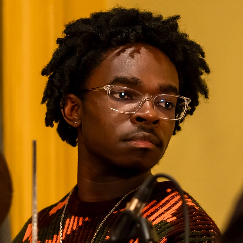 A young person with short, curly black hair and clear glasses looks to the side with a serious expression. They wear a patterned sweater in orange, black, and brown tones. The yellow background frames Koleby Boyston beside a blurred microphone in the foreground.