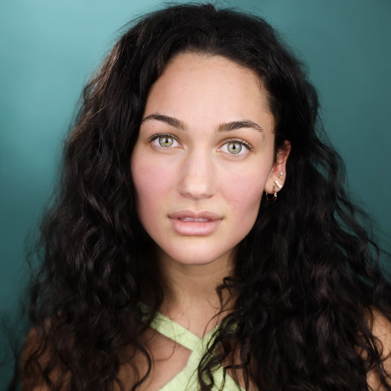 A woman with long, dark curly hair and green eyes looks directly at the camera. She has subtle makeup, gold earrings, and wears a light green, crisscross halter top. The softly blurred teal background complements Sydney Kollas’s striking gaze.