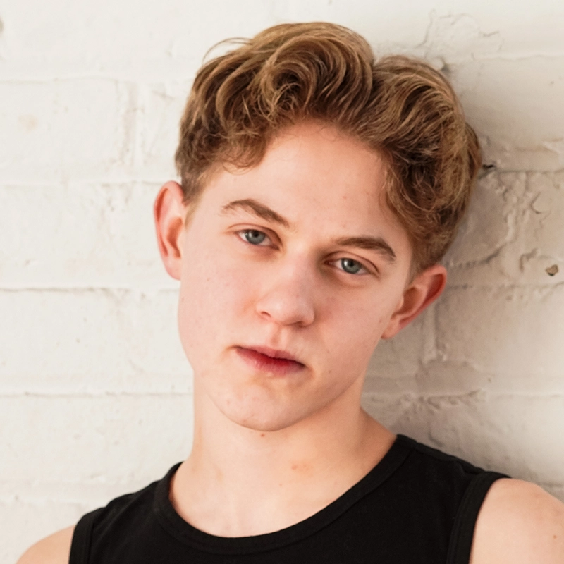 A young person with short, wavy light brown hair and fair skin stands against a white brick wall. They wear a black sleeveless top and have a neutral expression, with the head slightly tilted and eyes looking directly at the camera.