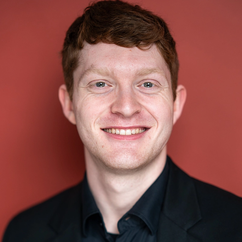 A young man with short, wavy red hair and fair skin smiles at the camera. He is wearing a black shirt and blazer. The background is a solid reddish color, adding a bold accent to Timothy Thomas’s confident presence.
