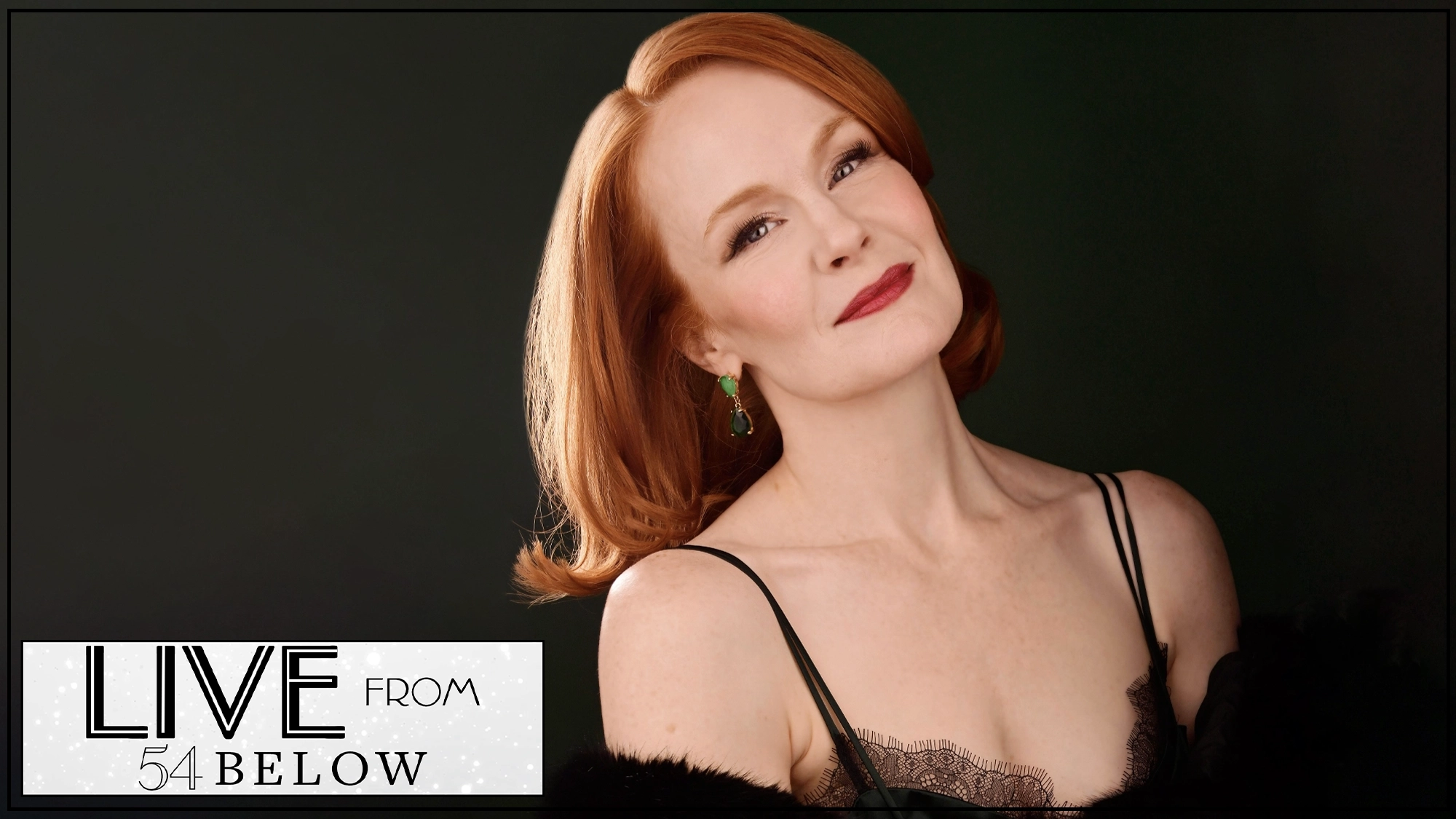 A woman with fair skin and red hair, resembling Kate Baldwin, smiles softly. She wears red lipstick, green earrings, and a black lace spaghetti strap top. The dark background features a 