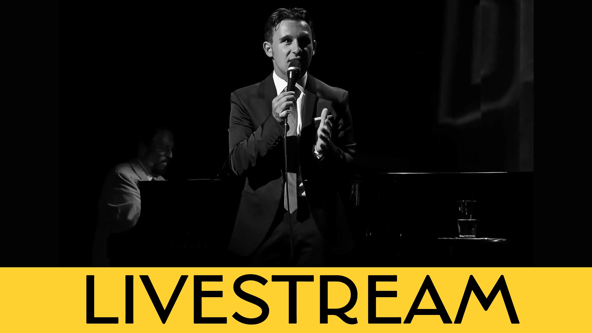 A man in a suit, Nicolas King, holds a microphone and speaks on stage before a dark backdrop with pianist Mike Renzi partially visible. At the bottom, a yellow banner displays the large black word 
