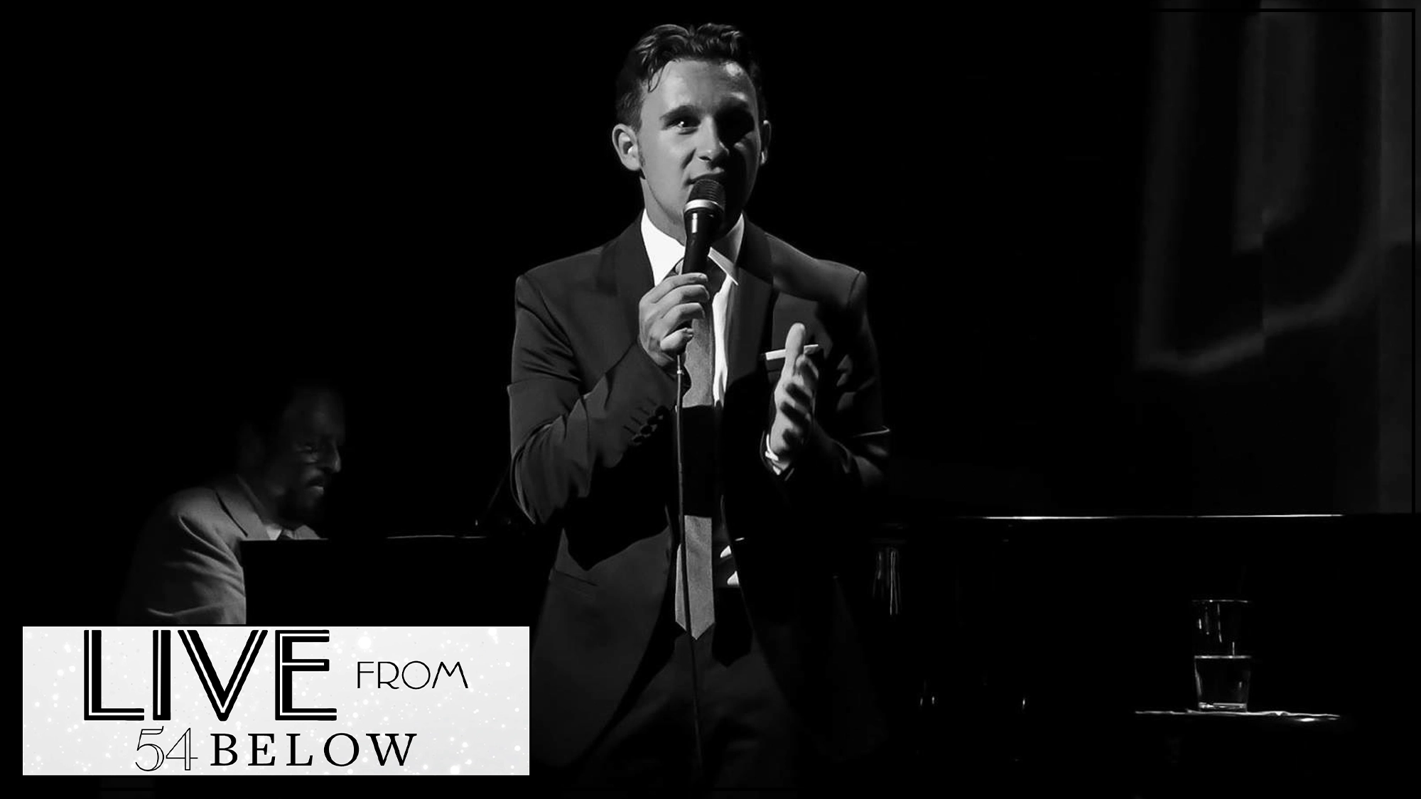 A man in a suit stands on stage, holding a microphone and speaking. He is lit by a spotlight against a dark background, with pianist Mike Renzi partially visible behind him. In the corner, a logo reads “LIVE FROM 54 BELOW - LIVESTREAM.”.