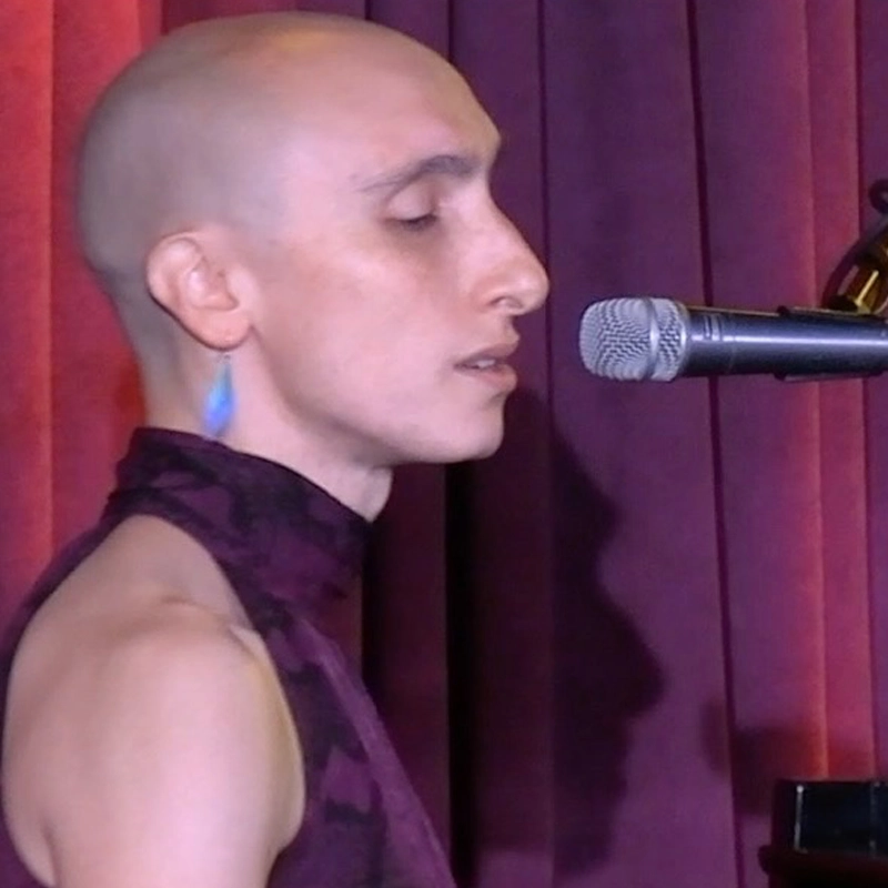 Elle Lagalante, with a shaved head, wears a sleeveless purple top and blue feather earring, singing or speaking into a microphone. She is shown in profile against red and purple curtains.