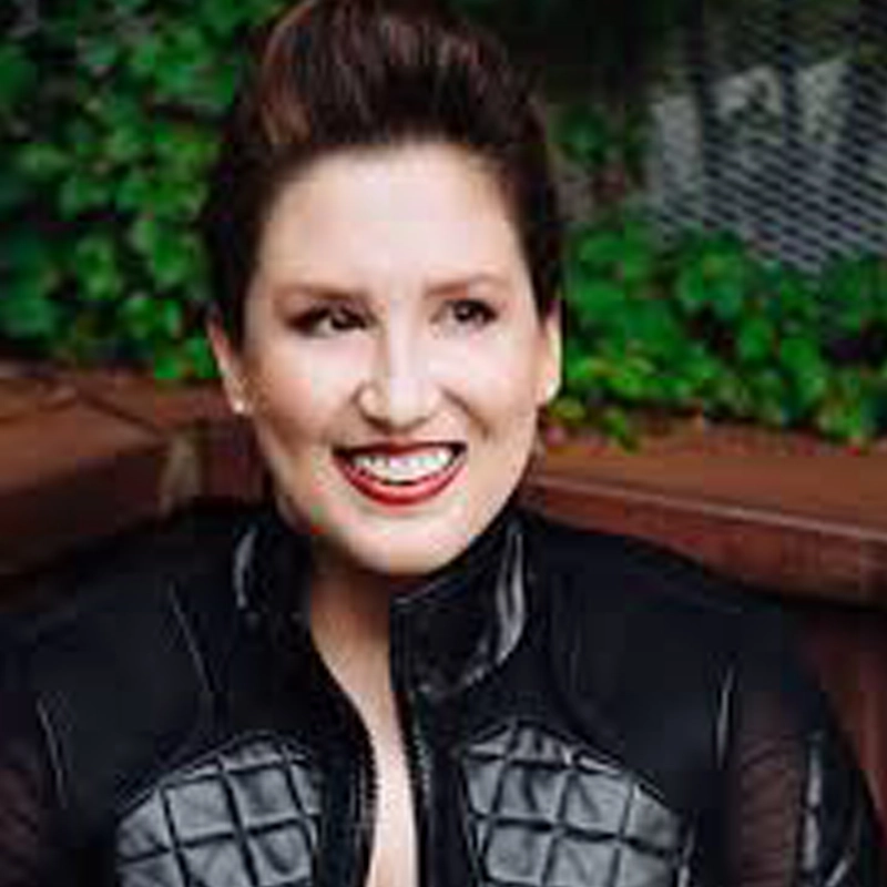 Alexandra Lang, a smiling woman with short, styled brown hair and bright red lipstick, sits outdoors in a black jacket with quilted panels. Green foliage and a wooden bench are behind her, as natural light creates a cheerful atmosphere.