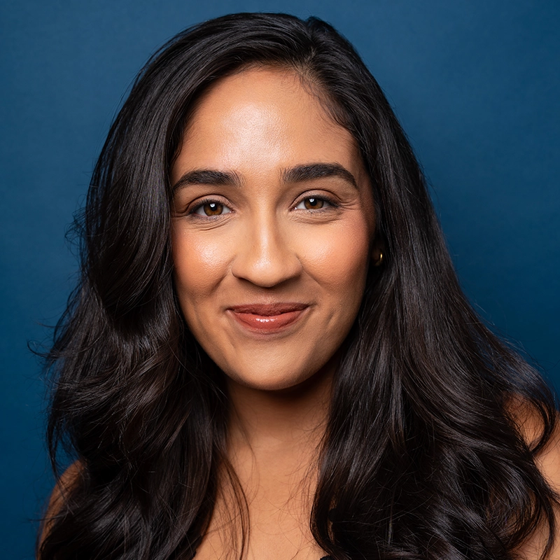 Lauren Marie Soto, with long, wavy dark hair, smiles softly at the camera. She has medium skin, brown eyes, and wears natural makeup. The solid dark blue background enhances her confident and approachable presence in this professional portrait.