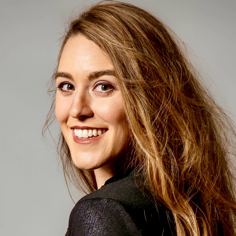 Chelsea LeValley, with long, wavy, light brown hair, smiles while looking over her shoulder. She has fair skin, wears a black top, and stands before a gray background. Warm lighting highlights her hair and friendly expression.