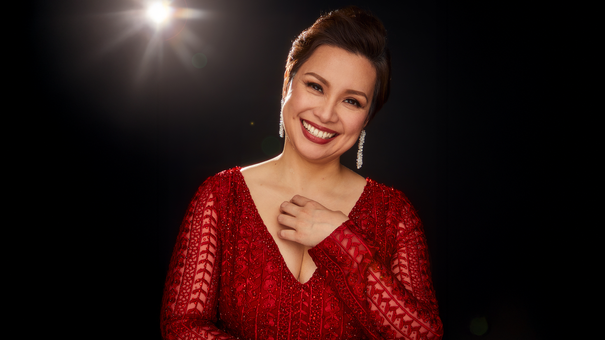 Lea Salonga - Site Image A woman with short dark hair smiles warmly, wearing a sparkling red dress with a deep V-neck and dangling earrings. She stands against a black background with a soft light shining behind her, creating a radiant and elegant atmosphere.