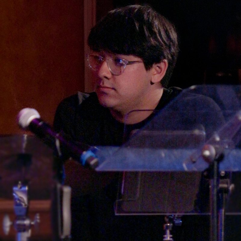 A young person with short dark hair and clear glasses sits behind a music stand, looking thoughtfully to the side. They wear a black shirt. A microphone and transparent barrier are visible in the foreground, suggesting a music or recording setting.