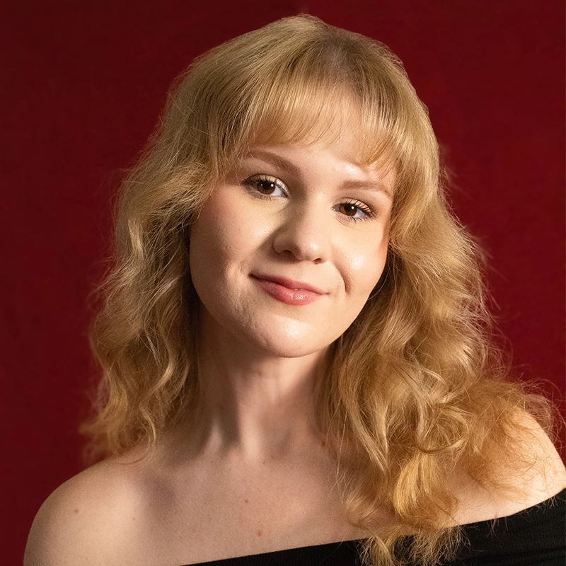 A young woman with wavy blonde hair and bangs smiles gently, facing forward. She wears an off-the-shoulder black top against a deep red background. The lighting softly highlights her clear skin and relaxed expression, reminiscent of the portrait style often favored by Timothy Thomas.