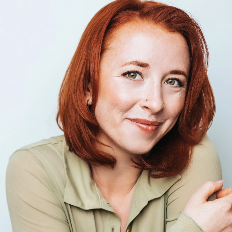 Allison Lian, a woman with straight, shoulder-length red hair and fair skin, smiles softly. She has green eyes, a small nose stud, and wears a sheer, light green blouse. Her arms are gently crossed against a pale background.