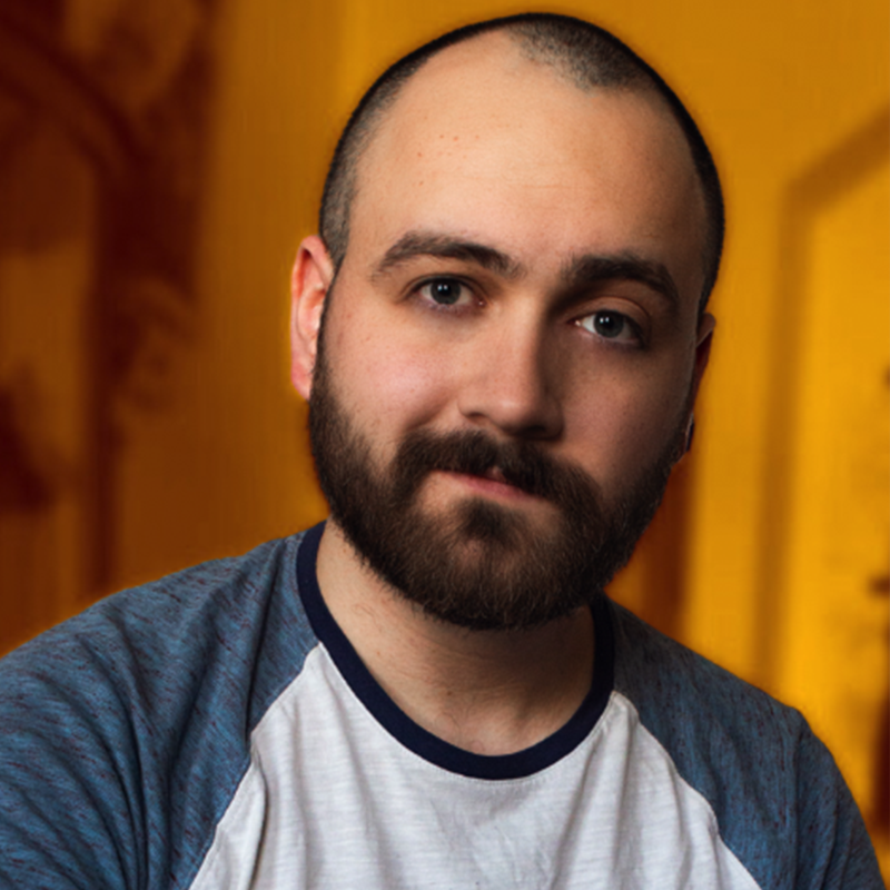A bearded man with a shaved head, wearing a blue and white raglan t-shirt, looks at the camera with a gentle expression. The background is warmly lit in orange and brown tones, softly blurred to draw focus to the man's face.