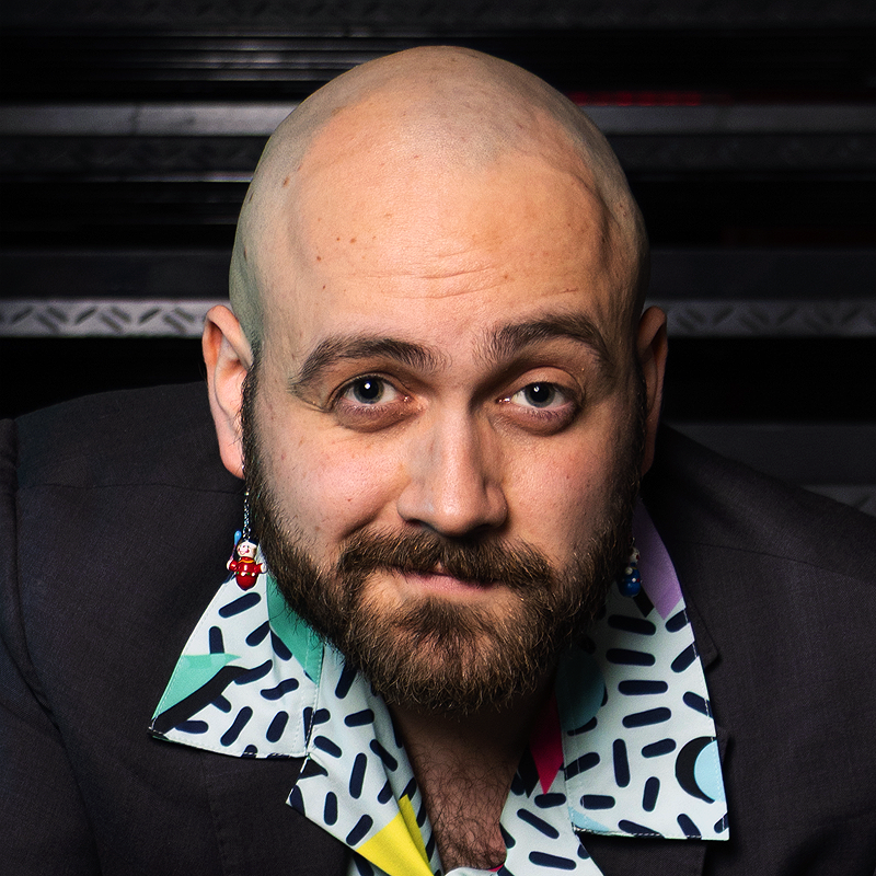 A bald man with a full beard looks at the camera, wearing a suit jacket over a colorful, patterned shirt. He has drop earrings shaped like red dice. The background is dark, with a metallic, industrial texture behind him.