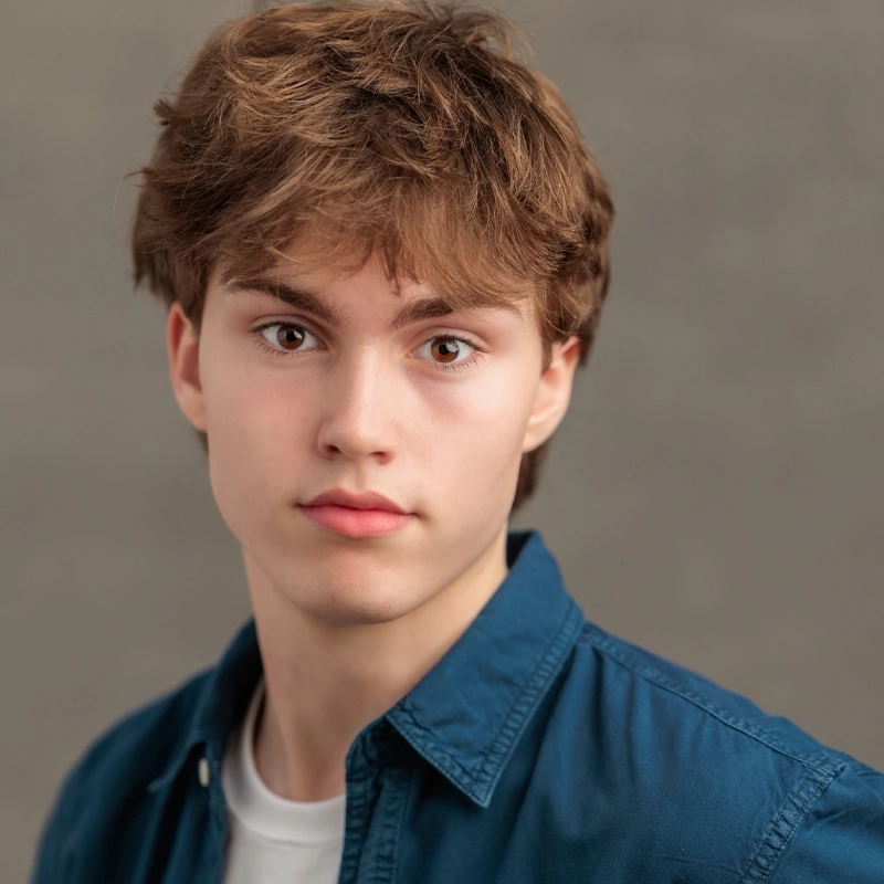 A young person with light skin and tousled brown hair looks directly at the camera with a neutral expression. Daniel Loughlin wears a blue collared shirt over a white t-shirt, set against a plain, softly blurred background in gray tones.