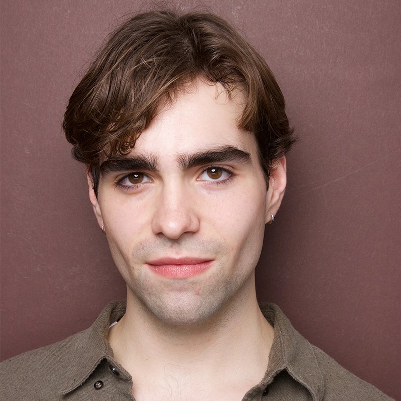A young person with fair skin and short, wavy brown hair stands against a plain mauve background. They wear a khaki-colored shirt and small hoop earrings, and have a neutral expression with a slight smile.
