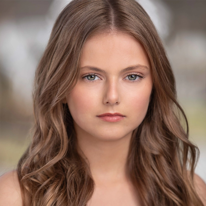 A young woman, Luli Mitchell, with long, wavy light brown hair and fair skin faces the camera. She has blue eyes, a neutral expression, and wears subtle makeup. The background is blurred, drawing focus to her face.
