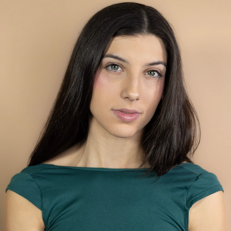 A young woman with straight, long dark brown hair poses against a peach background. She wears a green off-the-shoulder top and has light skin, neutral makeup, and a subtle gloss on her lips. She looks directly at the camera with a calm expression.