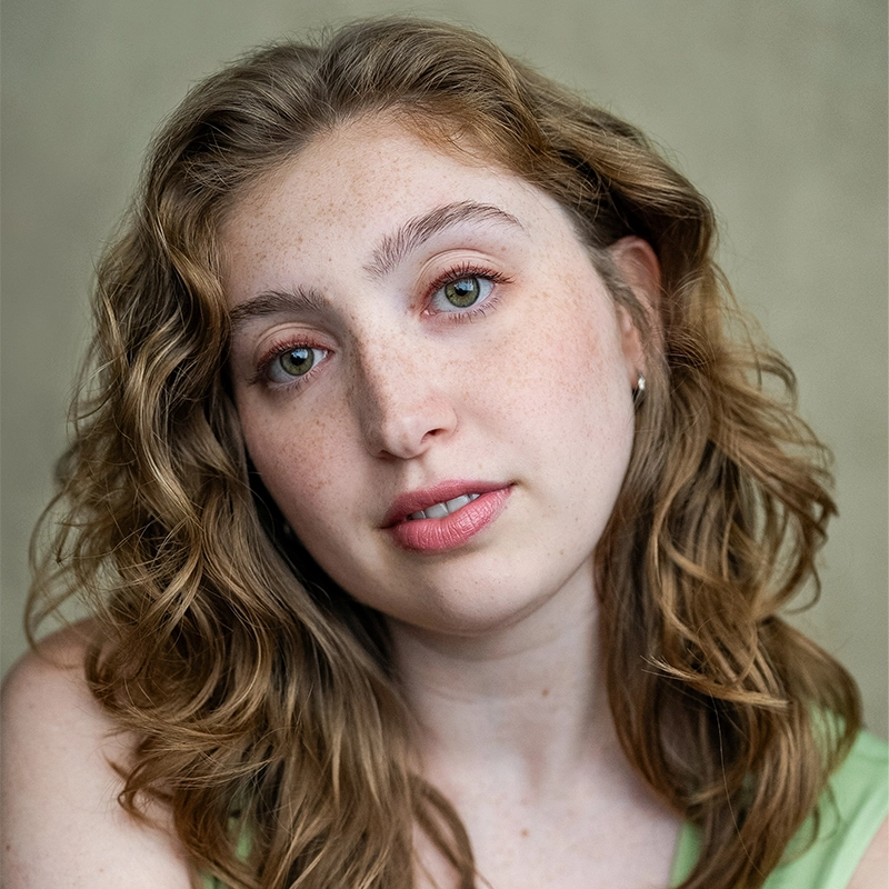 A young woman with wavy, light brown hair and green eyes looks softly at the camera. She has fair skin with freckles, lightly pink lips, and wears a pale green top—capturing the gentle style of Koleby Boyston. The background is muted beige.