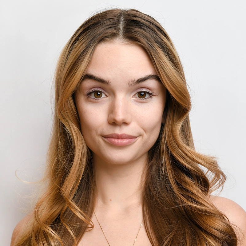 A young woman with long, wavy, light brown hair and fair skin looks directly at the camera. She has arched eyebrows, brown eyes, and a slight smile. The plain white background highlights Maggie Weller’s natural beauty.