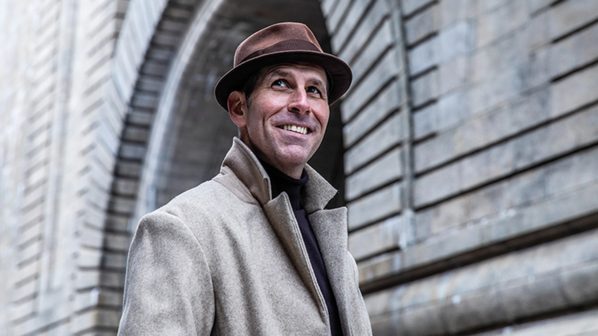 Marcus Goldhaber site image 26 A smiling man in a brown fedora and beige coat stands outside by a large stone archway. Reminiscent of Marcus Goldhaber’s