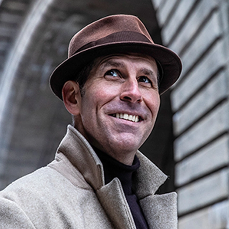 A smiling man wearing a brown fedora and a light-colored overcoat stands outdoors, looking up. He has light skin and is positioned in front of a stone archway with horizontal stone blocks, suggesting an urban setting.