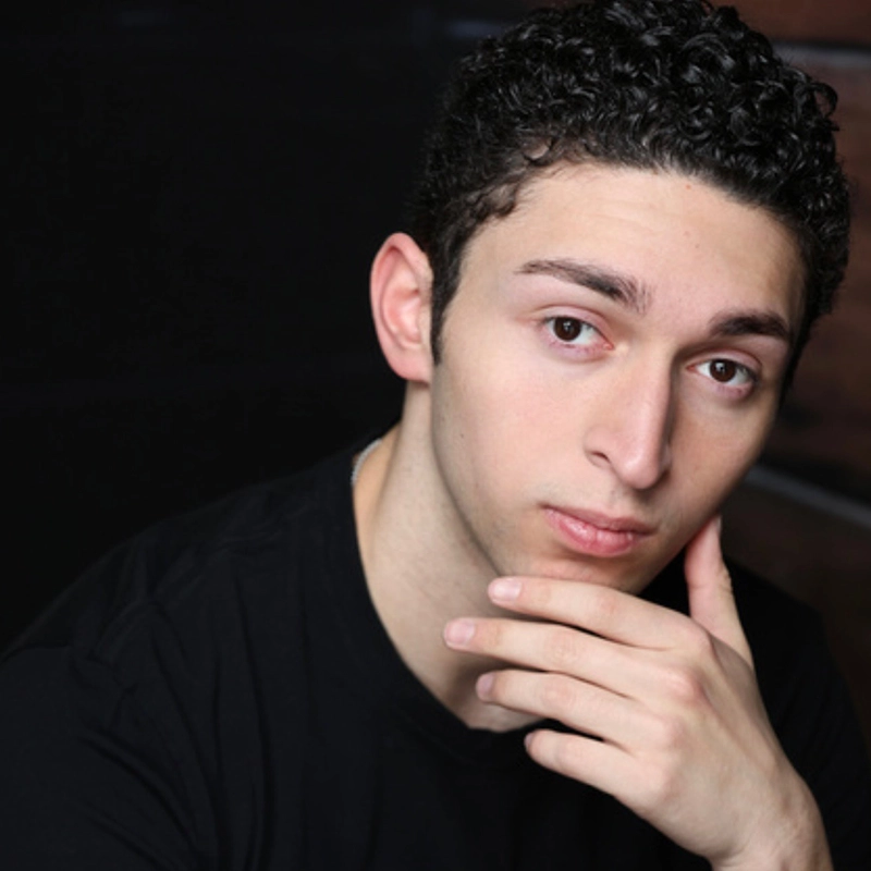A young person with short, curly dark hair and fair skin, resembling Jackie Winslow, looks into the camera with a neutral expression. They wear a black shirt and rest their chin on their right hand; the blurred dark background draws focus to their face.