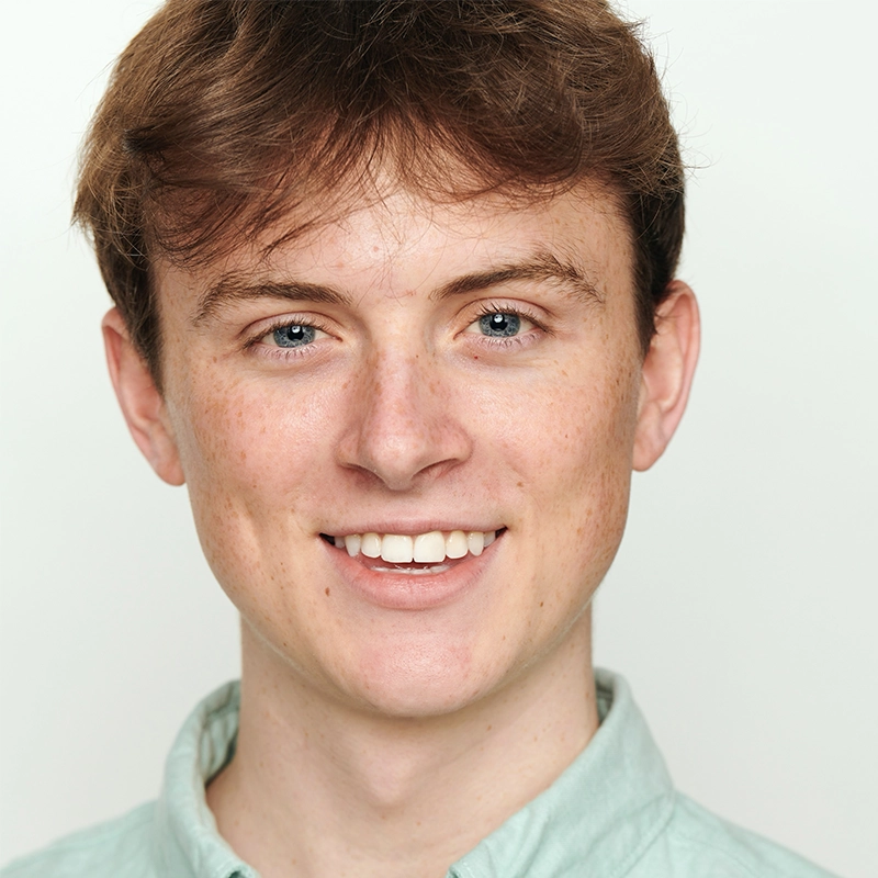 A young man with fair skin, short light brown hair, and blue eyes smiles at the camera. Wearing a light green collared shirt, he stands against a plain white background, his friendly expression reminiscent of Shayla Lopez’s approachable style.