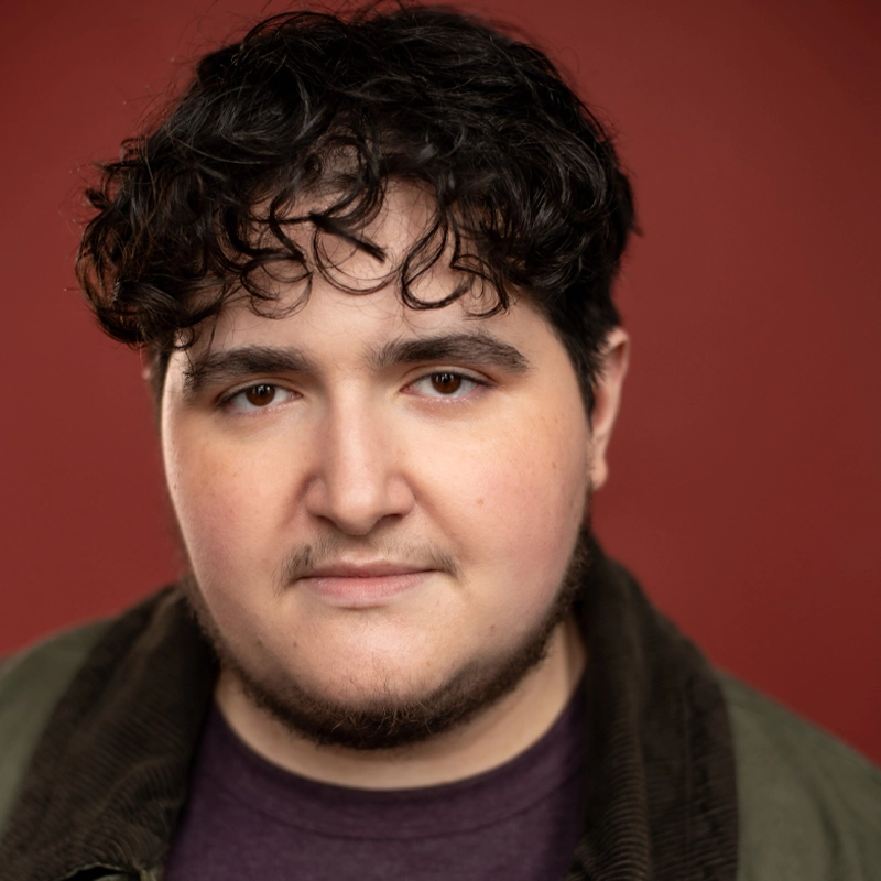 Emerson McAlister, with wavy dark hair and a short beard, looks into the camera with a neutral expression. He wears a dark jacket over a maroon shirt against a solid, muted red background with soft lighting.