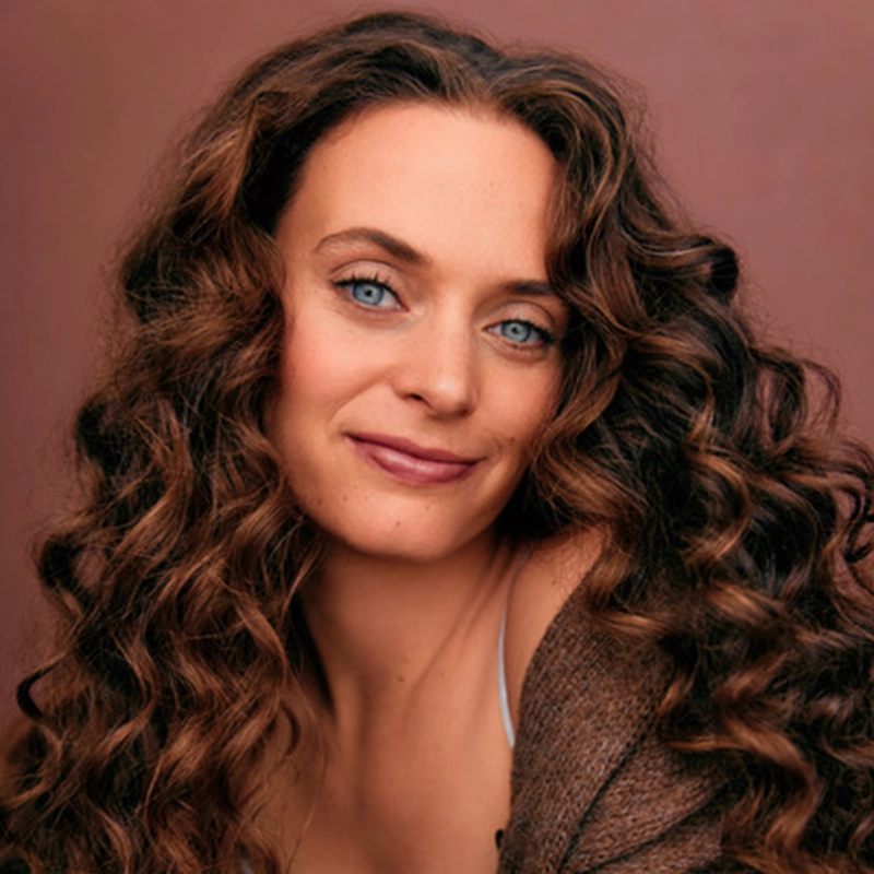 Jackie Winslow, with long, curly brown hair and blue eyes, smiles softly at the camera. She wears a brown sweater off one shoulder and sits against a warm, muted brown background. Her expression is calm and inviting.