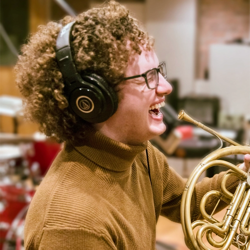 Zachary McIntyre, with curly hair, glasses, and large black headphones, smiles widely while holding a French horn. He wears a brown turtleneck sweater, and studio equipment including a drum set is visible in the blurred background.