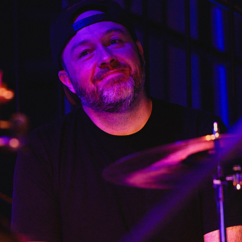 A bearded man in a black T-shirt and backward baseball cap, identified as Chris McWilliams, sits at a drum set. Purple and blue stage lights illuminate his face as he looks up with a slight smile. Drum cymbals and stands are visible in the foreground.