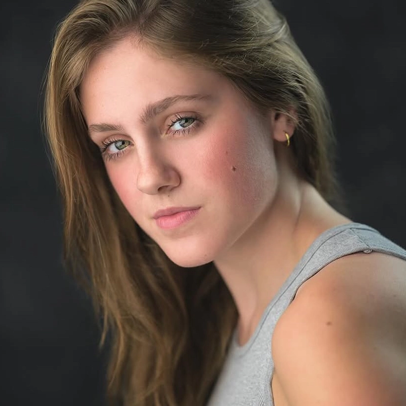 Sophia Leverett, a young woman with fair skin, long light brown hair, and blue eyes, gazes confidently at the camera. She wears a light gray sleeveless top and a small gold hoop earring. The dark, blurred background highlights her face.