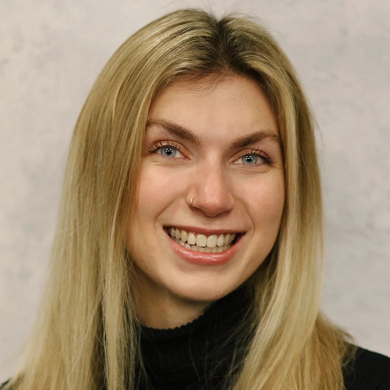 A young woman with long, straight blonde hair smiles at the camera. She has light skin, blue eyes, a gold nose ring, and is wearing a black turtleneck sweater. The background is neutral and softly textured.