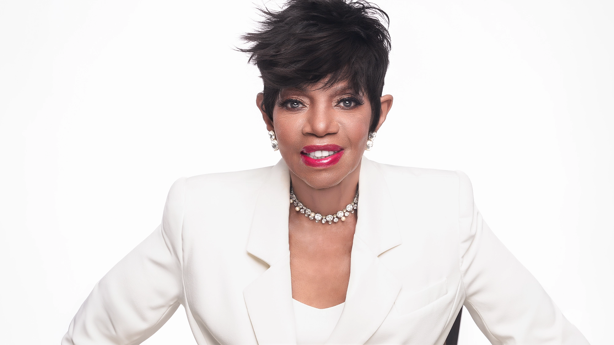 A woman with short, styled black hair wears a white blazer, pearl necklace, and pearl earrings. She is smiling with red lipstick against a plain white background, radiating the elegance of a Broadway star—reminiscent of Melba Moore in From Broadway With Love.