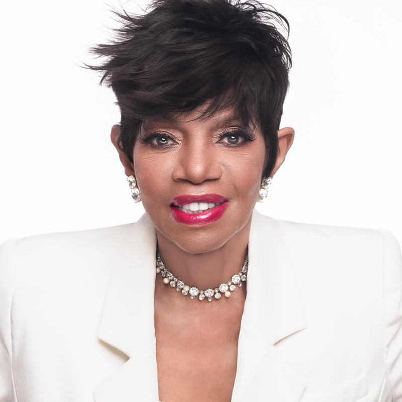 A woman with short, stylish black hair wears a white blazer, sparkling stud earrings, and a jeweled choker necklace. She smiles, showing bright red lipstick, against a plain white background. Her makeup is elegant and her look polished.
