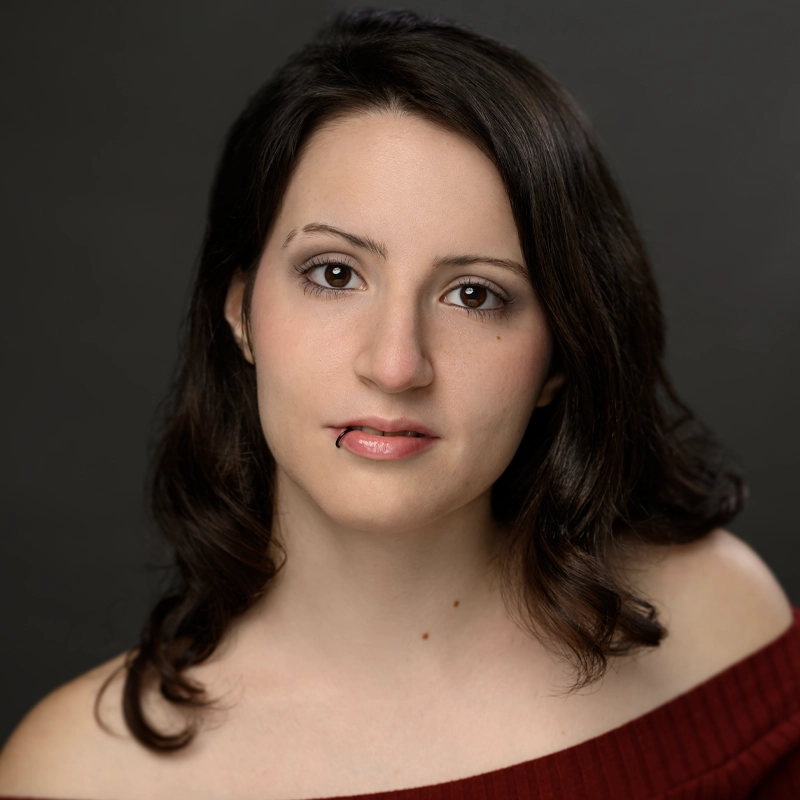Celeste Meola poses against a dark background, her shoulder-length dark brown hair framing her face. She wears an off-the-shoulder maroon top and a subtle lip piercing, with natural makeup highlighting her eyes and lips.