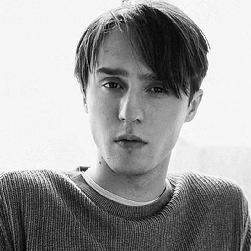 A young person with straight, dark hair parted to the side gazes seriously at the camera. They wear a textured sweater over a shirt. The background is bright and neutral, and the image is in black and white.