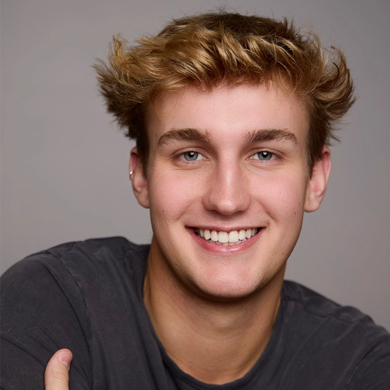 A young man with tousled blond hair and blue eyes smiles at the camera. He wears a dark grey t-shirt and sits against a plain, light grey background. His relaxed pose exudes the approachable style often captured by Alice K. Tinari.