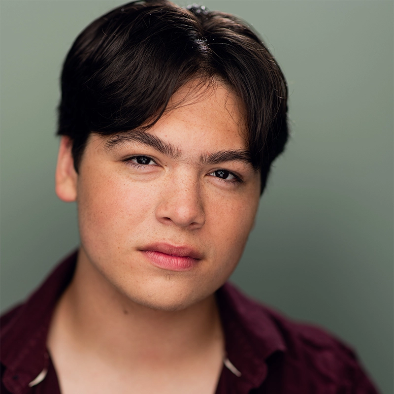 Shayla Lopez, a young person with short dark hair and fair skin, gazes directly at the camera with a neutral expression. Wearing a burgundy collared shirt over a light shirt, she stands against a softly blurred green background.