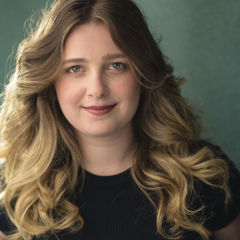 Lyndsey Minera, a young woman with wavy, shoulder-length blonde hair and fair skin, smiles softly. She is wearing a black top and stands before a muted green background, her expression calm and approachable in the soft, even lighting.