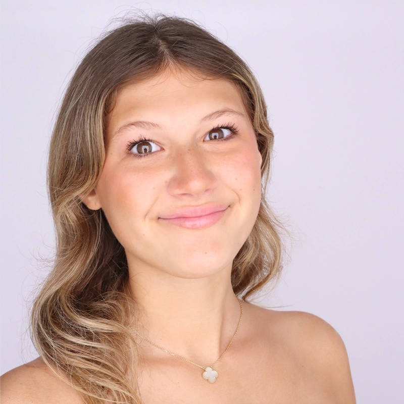 A young woman with wavy, light brown hair smiles gently at the camera. She is wearing a gold necklace with a four-leaf clover pendant. Her shoulders are bare, and the background is plain white.