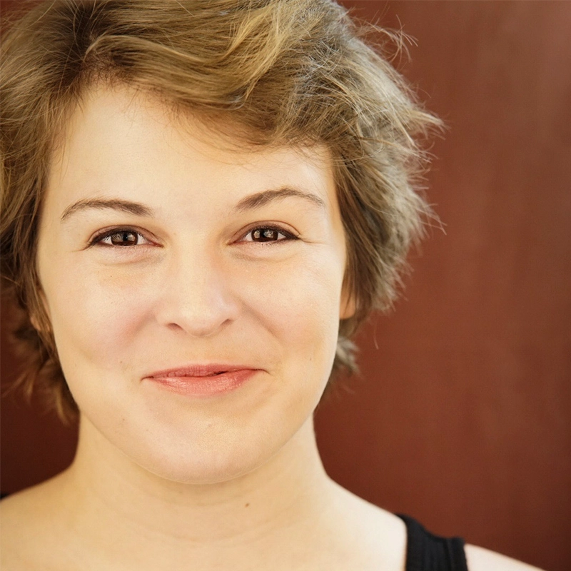 Maggie Weller, with short, tousled light brown hair, smiles gently at the camera. She wears a black sleeveless top and stands against a plain reddish-brown background. Soft lighting highlights her natural features.