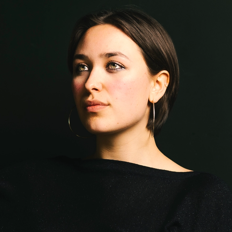A young woman with short brown hair and hoop earrings looks slightly upward. She wears a black top and sits against a dark background, her face softly lit, highlighting her thoughtful expression.