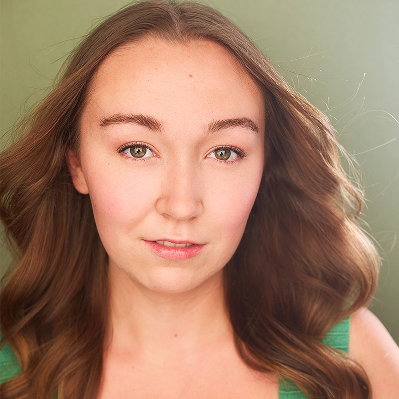 Luli Mitchell, a young woman with long, wavy brown hair and light skin, looks directly at the camera while wearing a green sleeveless top. The background is a muted green. Her expression is calm and confident, enhanced by natural makeup and soft lighting that gently illuminates her face.