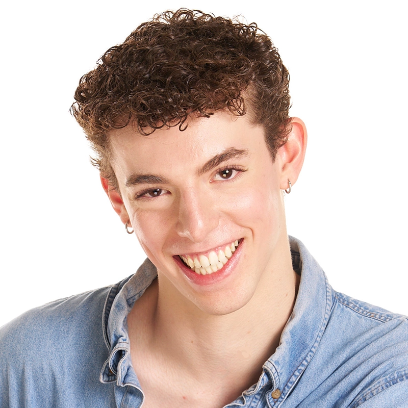 A young person with short, curly brown hair and fair skin smiles widely. Maggie Weller wears small hoop earrings and a light blue denim shirt with the collar casually open. The plain white background highlights their cheerful expression.