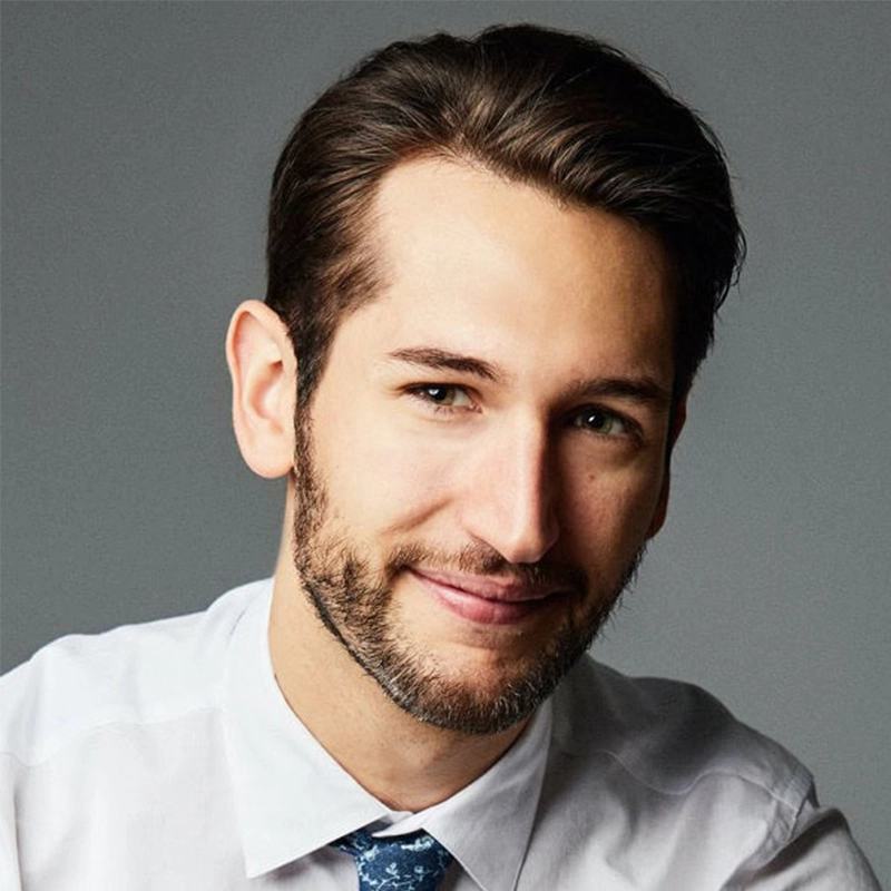 A man with short dark hair and a trimmed beard smiles gently. He is wearing a white dress shirt and a blue tie with a subtle pattern, reminiscent of Amanda Quaid's classic style. The plain grey background and soft lighting highlight his friendly expression.
